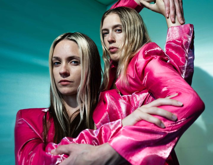 2 female performers wearing electric pink coats dancing against a teal green background