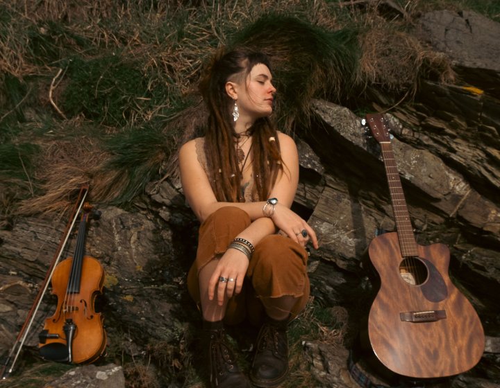 A musician sat on a rock with a violin on the left and an acoustic guitar on the right