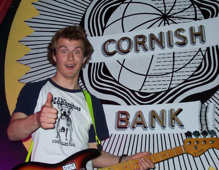 Image of a person holding a guitar in front of the colourful Cornish Bank logo painted on the wall