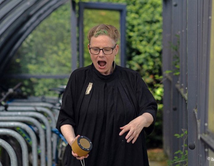 A performer located in a bike shed wearing black clothing holding a wireless speaker
