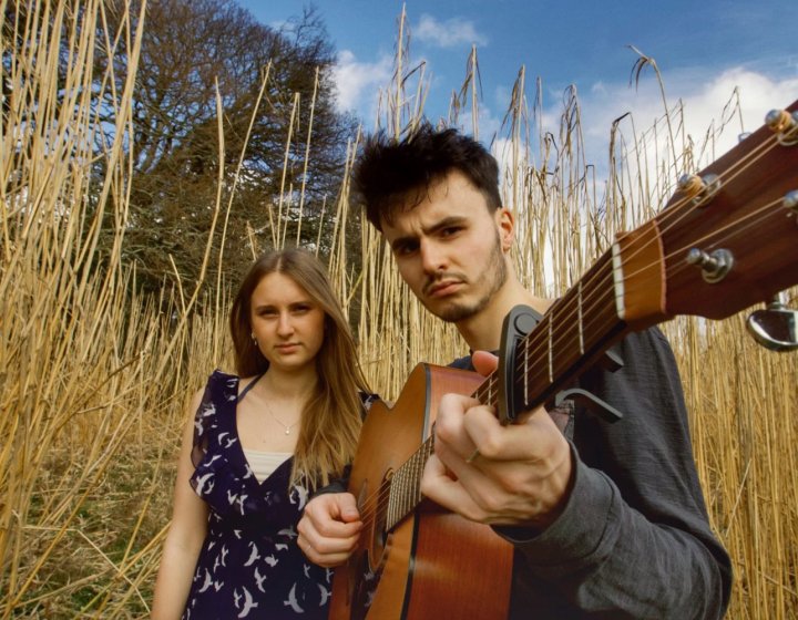 Two performers outside against a long grass background. The performer in the front of the image is holding a guitar