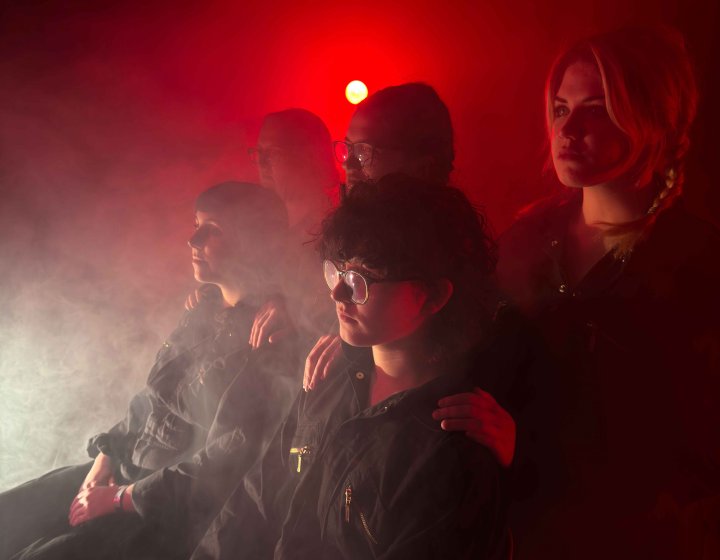 4 people seated and standing looking away from the camera on stage with a red light behind them with lots of smoke