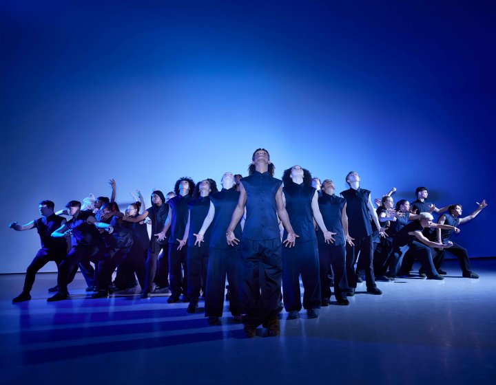A large ensemble of youth dancers in dark clothing against a striking blue backdrop