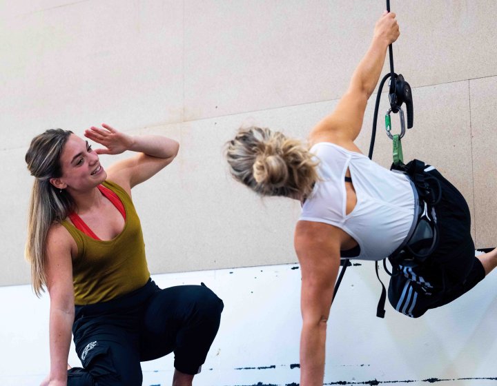 2 performers up against a wall, 1 of them is being supported by an aerial dance harness