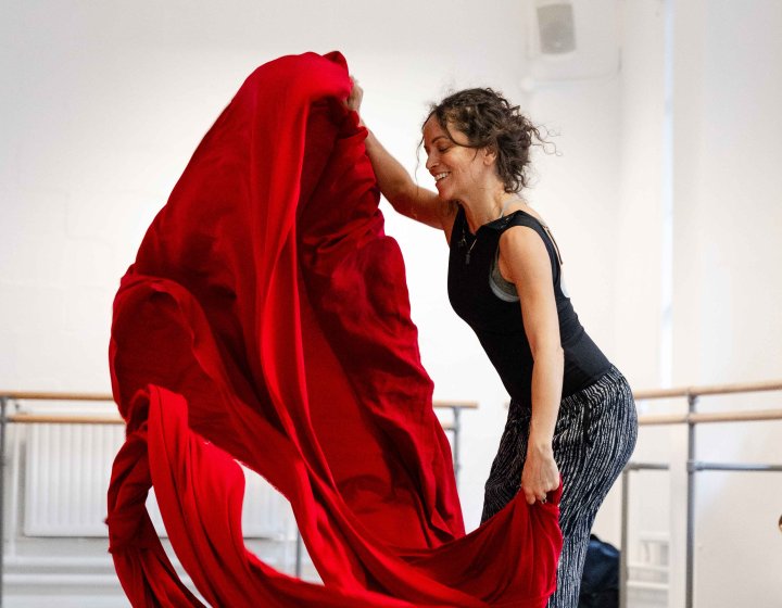 A dancer wearing dark clothing with a large piece of red fabric in a white dance studio