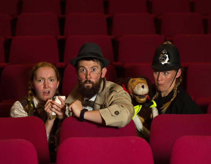 3 people sat on red cinema style chairs dressed up as police officers and 1 person has a puppet