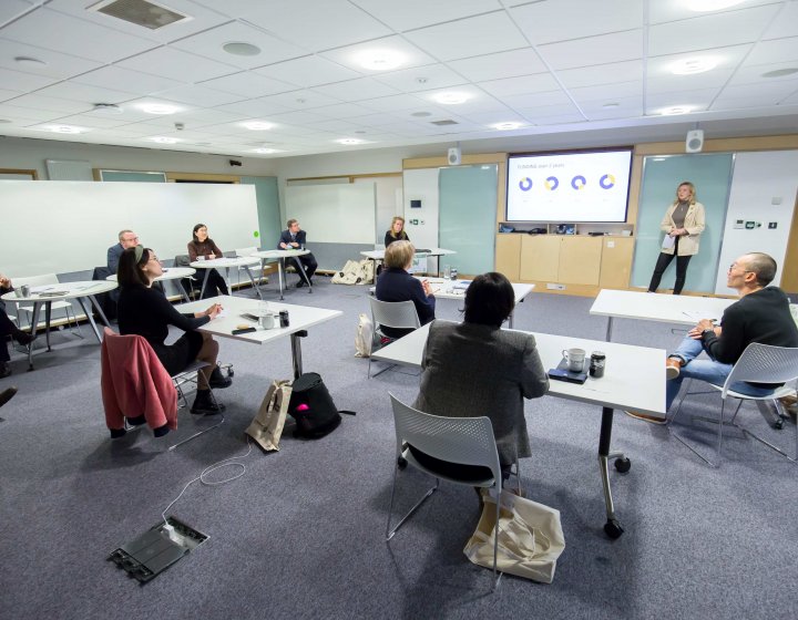 A student stands in front of a television screen and presents their business pitch to a panel of experts