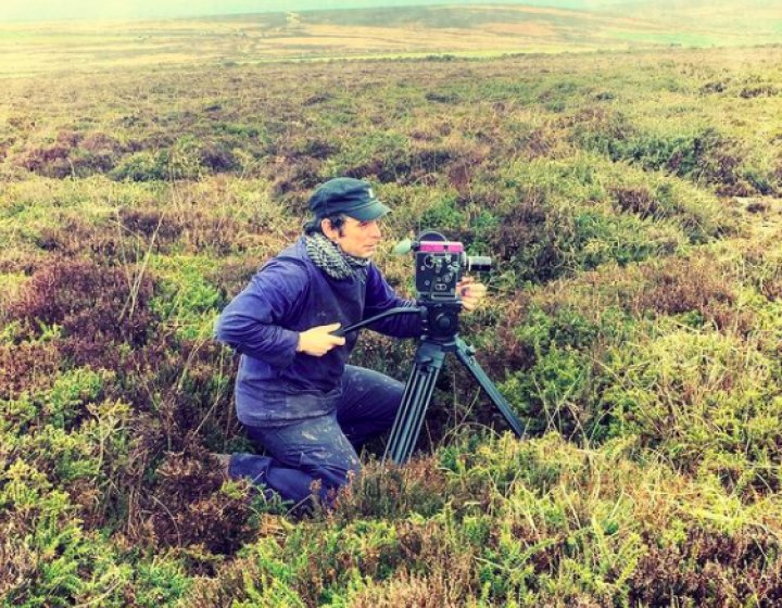 A filmmaker crouches behind his camera amongst shrubbery 