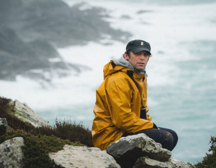 A man in a yellow coat sits atop a cliff, the ocean set behind him