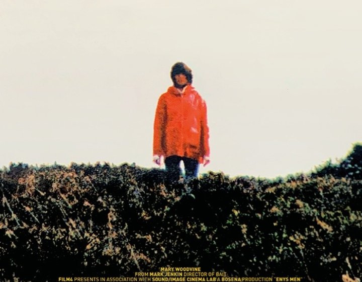 Film poster for ENYS MEN shows woman wearing jeans and red coat standing amongst yellow gorse. Text reads: From the director of BAIT, ENYS MEN, a film by Mark Jenkin