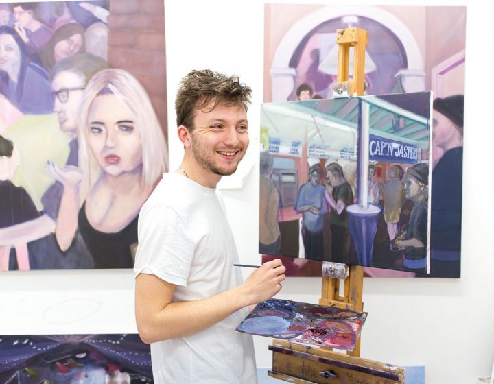 Student stood in front of paintings, smiling and holding paintbrush