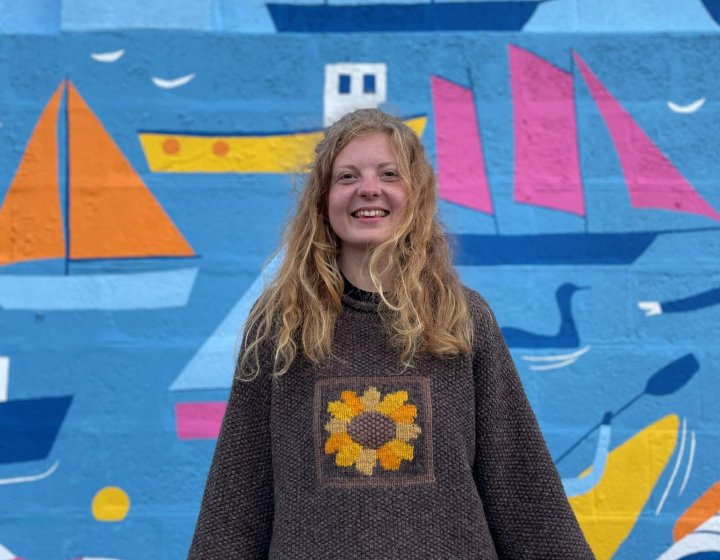 A girl wearing a brown jumper standing in front of a mural with boats