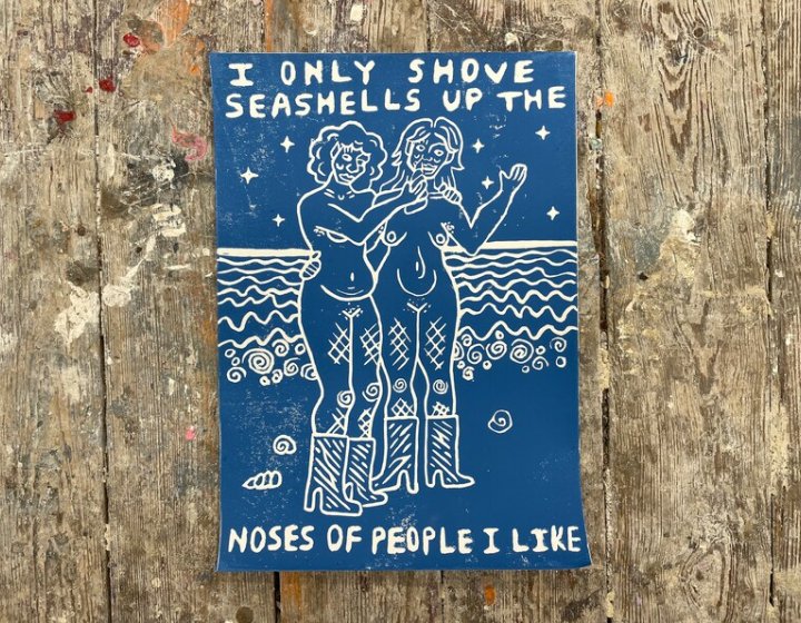 Blue and white lino print of two naked women on a beach. Words in white read: I only shove seashells up the noses of people I like.
