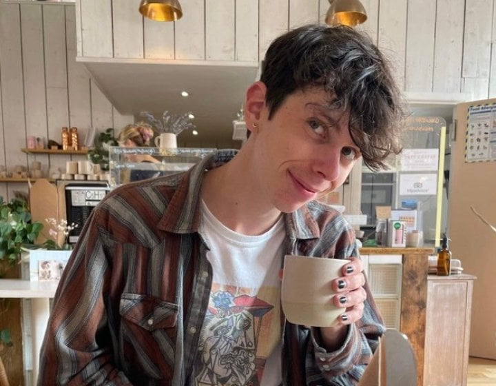 Photo of student Nathan Copeland wearing a graphic tee and checked shirt posing with a cup of coffee in a cafe.
