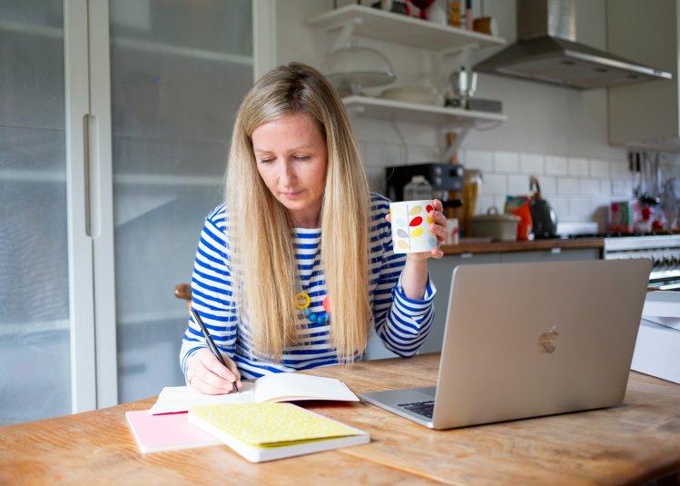 Woman at laptop writing script on paper, holding mug