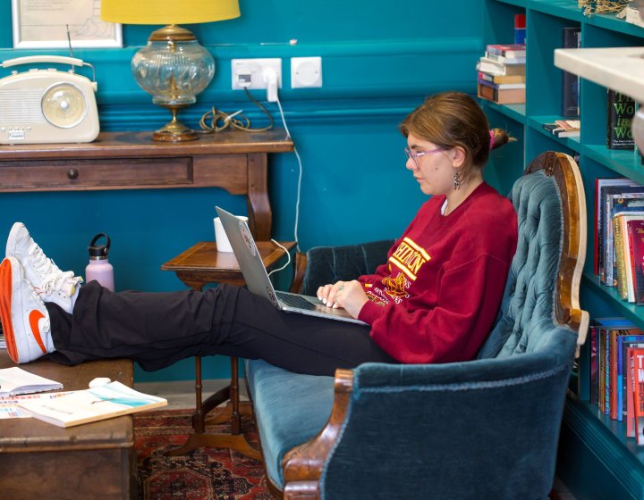 Girl wearing red jumper and pink glasses sits on a blue sofa with her feet up on a coffee table while working on a laptop