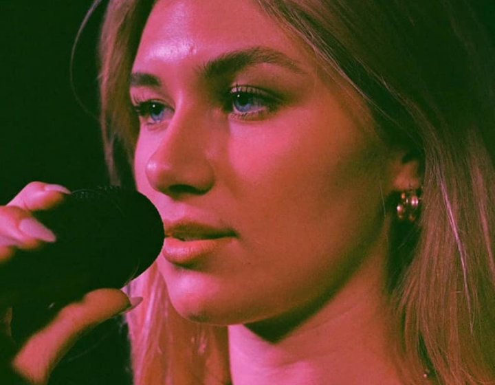 A close up of a woman singing into a microphone. She wears a gold necklace and is bathed in pink light