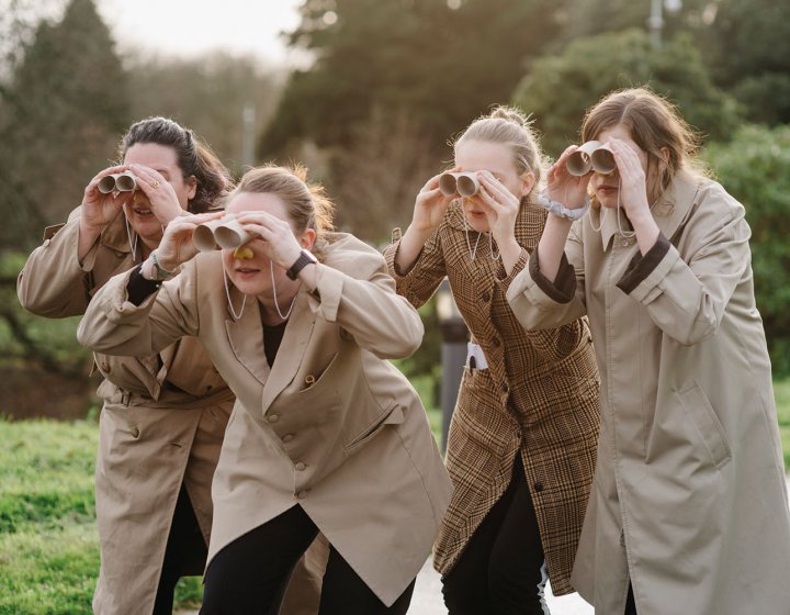 Student actors wear beige jackets and holding toilet rolls as binoculars
