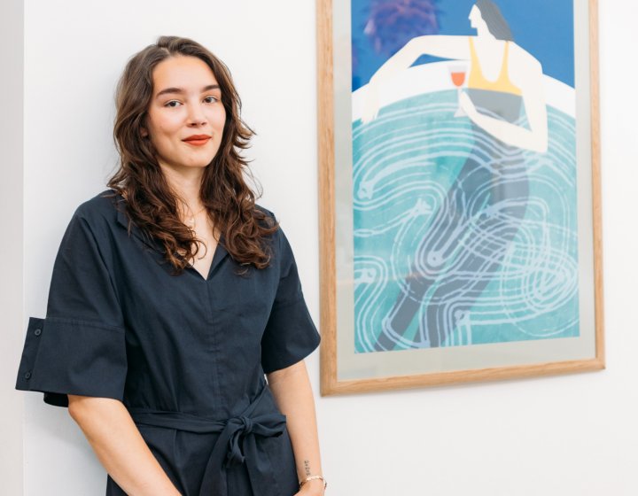 A young woman with dark hair standing next to a framed artwork of a woman drinking a glass of wine in a pool