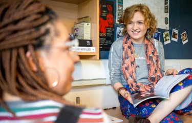 Student with book open on her lap chatting to another student in their room.