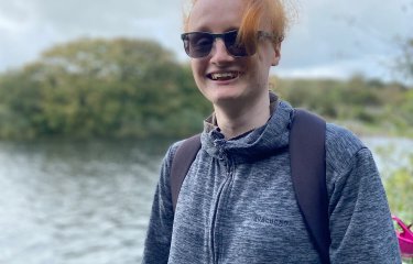 A Falmouth University students next to a water and trees, wearing a hoodie and sunglasses