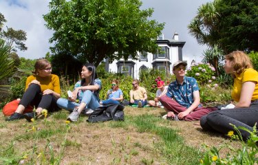 Students sat on the grass at Falmouth campus in front of an old black and shite Victorian building.
