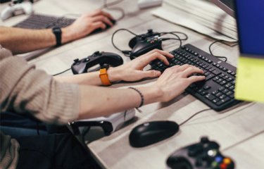 Hands typing on a keyboard with playstation controllers on the table