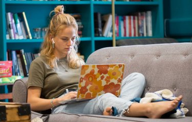 A student reclines on a sofa, working on her laptop