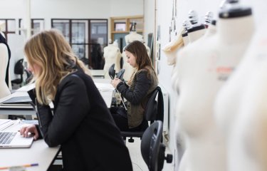 Two students in fashion studio, one on laptop and one knitting.