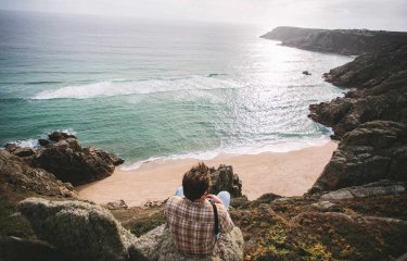 A Falmouth University student sitting on a cliff top overlooking a beach and sea