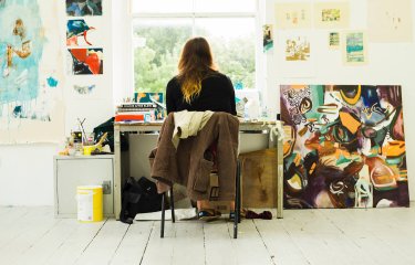 Back of student sitting at desk in front of window surrounded by artwork and paintings.