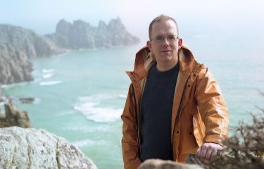 Writer, Wyl Menmuir, standing on cliff with sea view in the background.