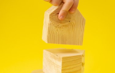 A set of three wooden blocks against a yellow background with a hand in shot holding one block 