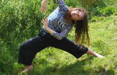 A person dancing outside on green grass with a bush in the background
