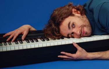 A person laying on top of a piano keyboard against a royal blue backdrop