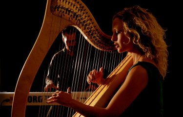 A close image of a harpist holding and playing the instrument with a person in the background playing an electric piano