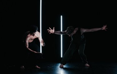 Two dancers on stage in very dim light performing around two white strip lights