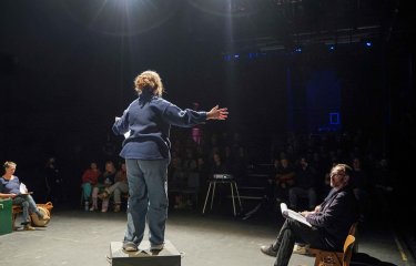 Performer with their back to the camera on stage under a spotlight performing to an audience