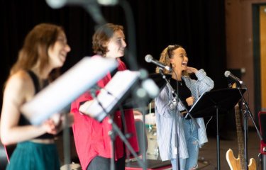 Three performers holding sheet music singing into microphones on stage