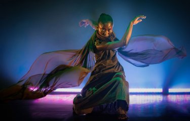A dancer wearer colourful costume with a pink/blue uplighter behind them