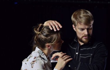 Two performers dancing on in close contact against a dark black backdrop