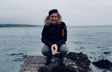 A male student crouching on rocks with sea in the background