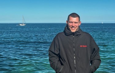 Portrait shot of Ben Penrose wearing black beach robe with red Zen Beach logo, with blue sea in the background