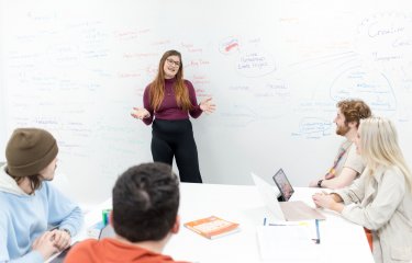 A student leads a discussion, with a large whiteboard full of notes behind her.