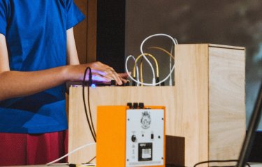 A young person in a blue t-shirt with their hand on a synth