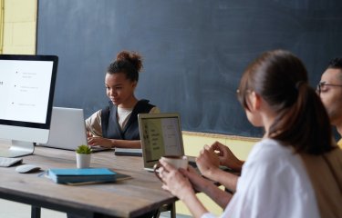 Three people studying at laptops on a table with a notebook and plant