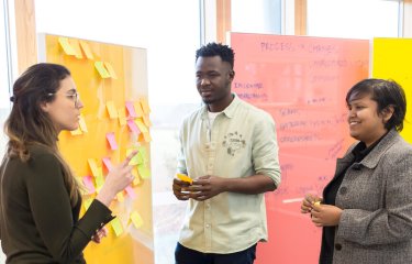 Three people standing in front of a yellow board covered in post-it notes