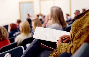 A person sitting in a lecture theatre with a notebook and pen in hand