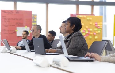 A group of people sat with laptops on a table, board of post-it notes behind them