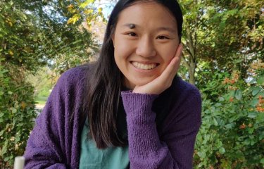 Female student wearing purple jumper 4smiling while sitting at wooden bench with leafy trees in the background.